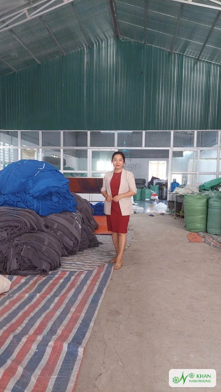 Lê Thị Nhung – Người Sáng Lập, Giám Đốc (Ceo) Khăn Nam Phong Lê Thị Nhung đứng cạnh các kiện khăn mộc màu xanh và xám trong kho nhà máy