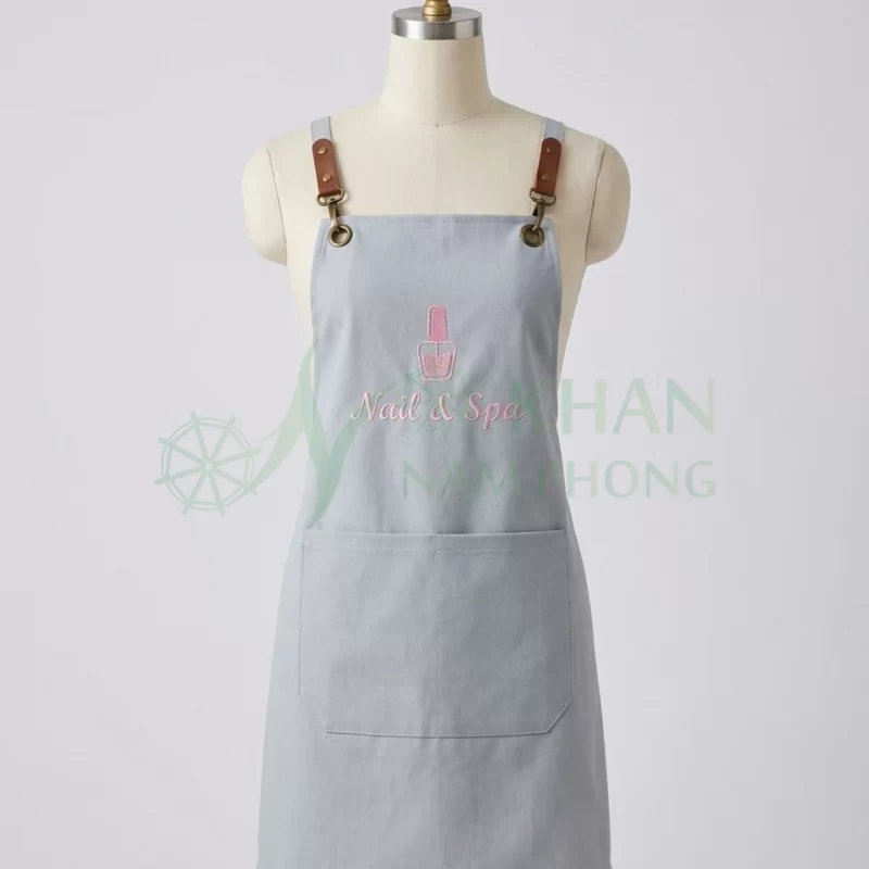 Tạp dề nail màu xám xanh mặt trước: yếm xám xanh, hai quai có khóa chỉnh trên vai, logo thêu trước ngực, túi ngang bụng